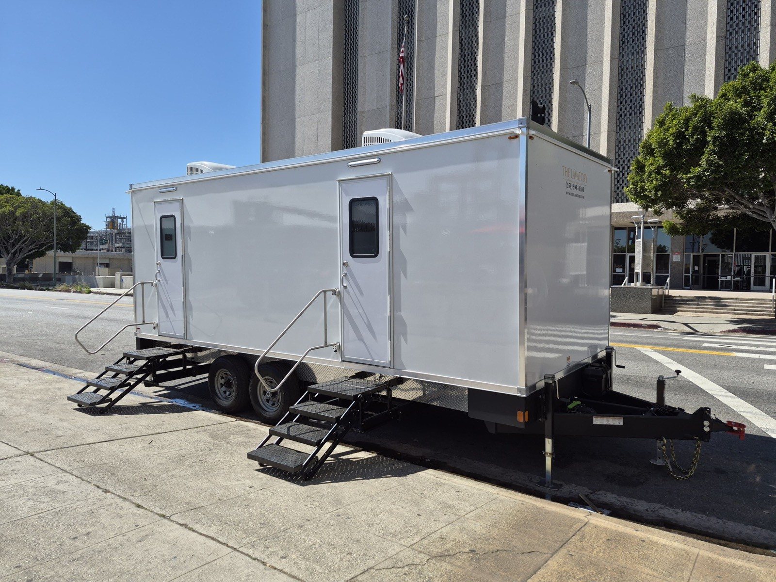 A Lavatory restroom trailer parked along a city curb in front of a tall building