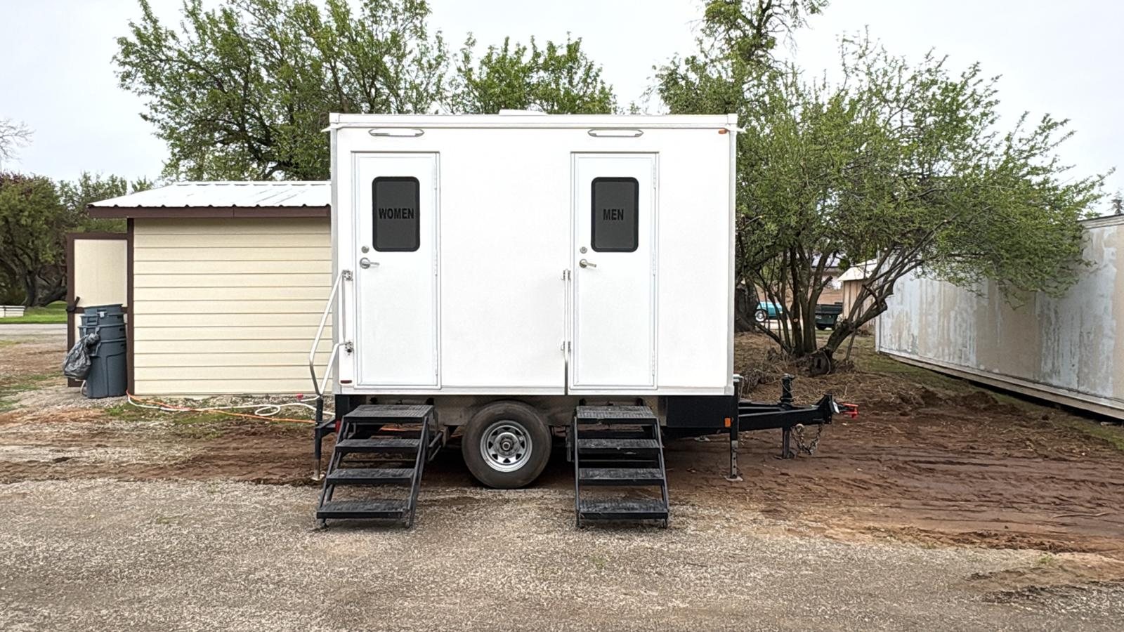 A Lavatory 2-Station restroom trailer on a dirt work site beside a small shed and a row of trees