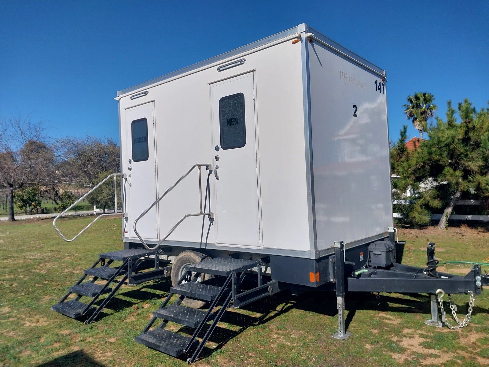 A Lavatory 2-Station restroom trailer on a grass lawn with a palm tree and pines behind it