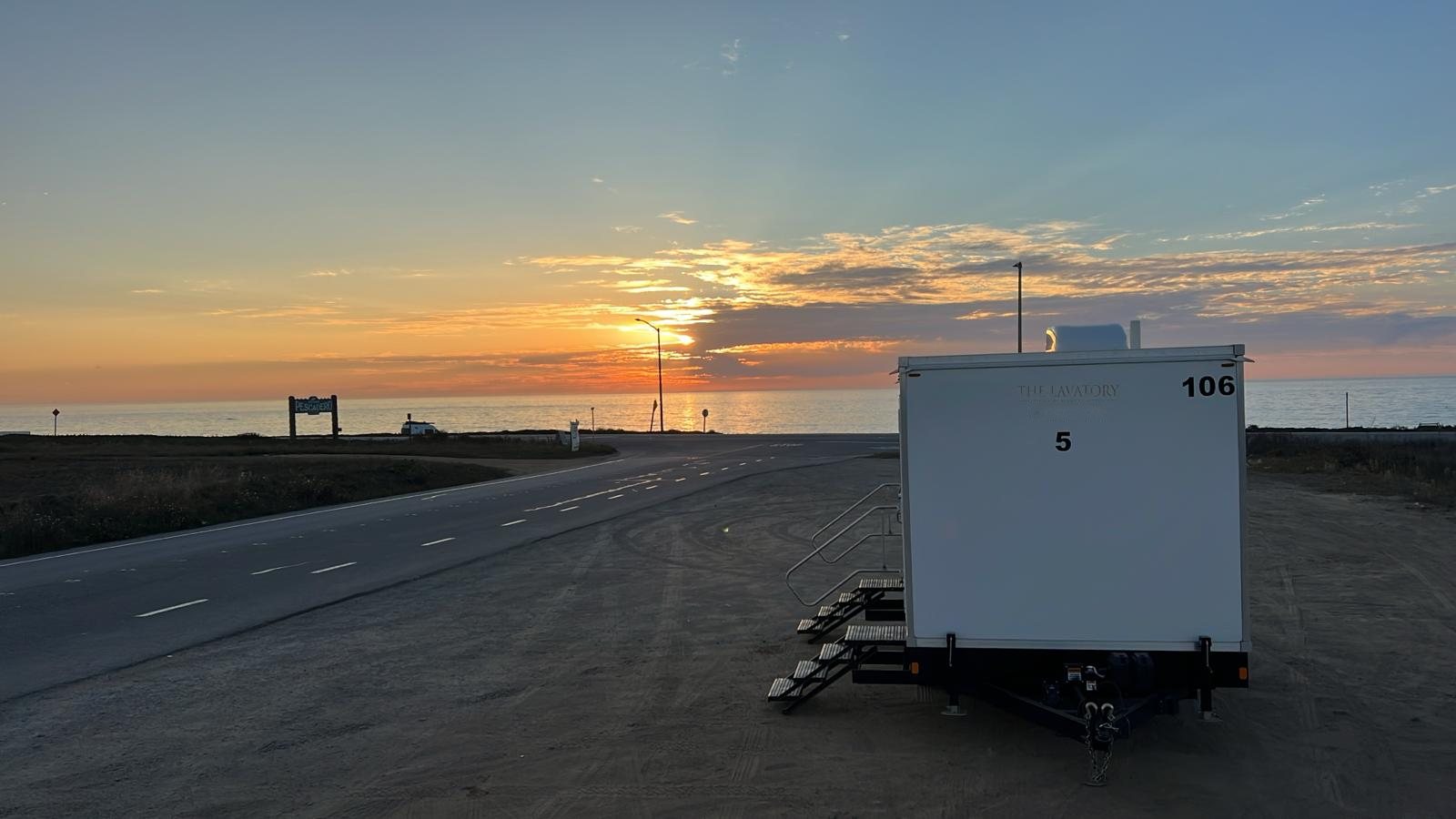 A Lavatory 5-Station luxury restroom trailer parked on a coastal lot at sunset, with the ocean behind it