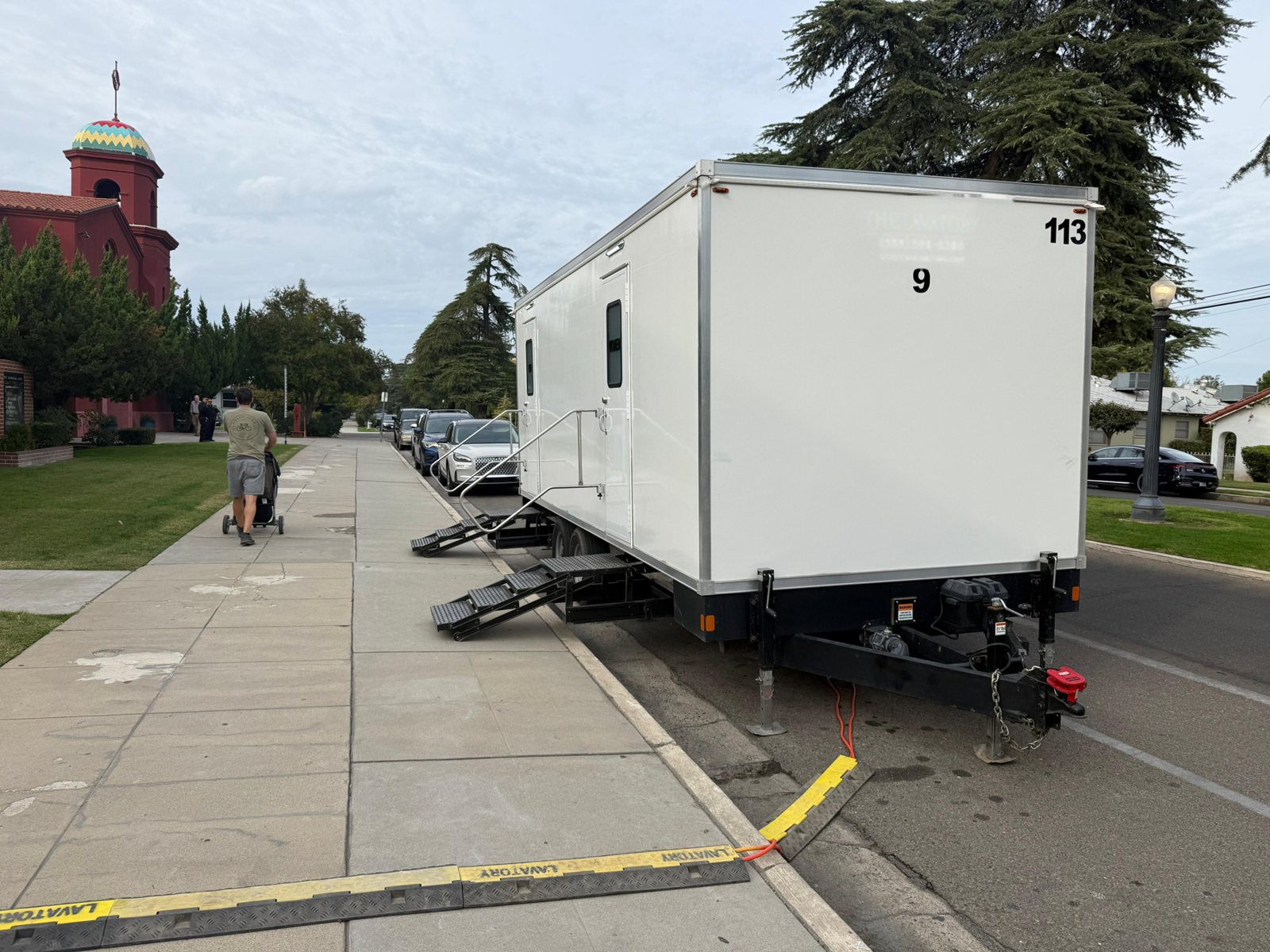 A Lavatory 9-Station restroom trailer parked along the curb beside a domed church, with a man pushing a stroller down the sidewalk