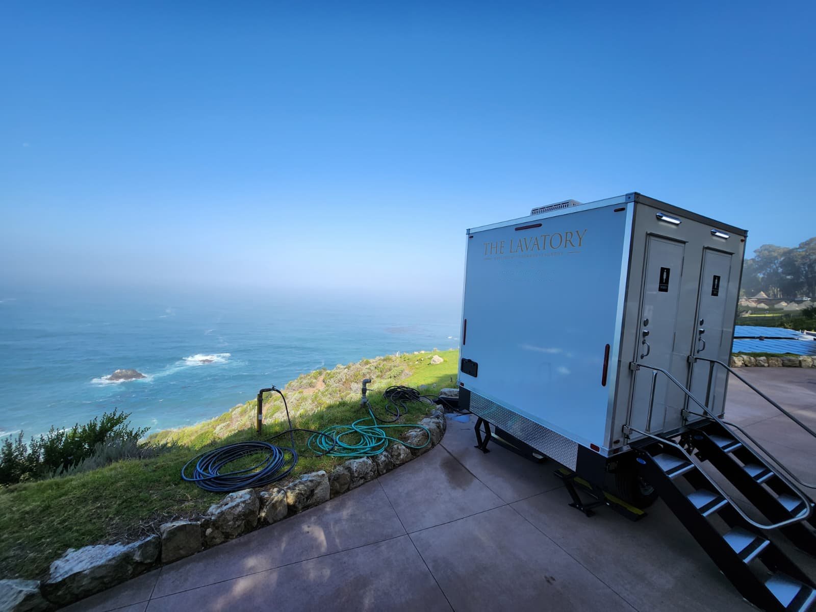 A two station Lavatory restroom trailer set on a patio above the ocean at a coastal home, with garden hoses coiled on the lawn