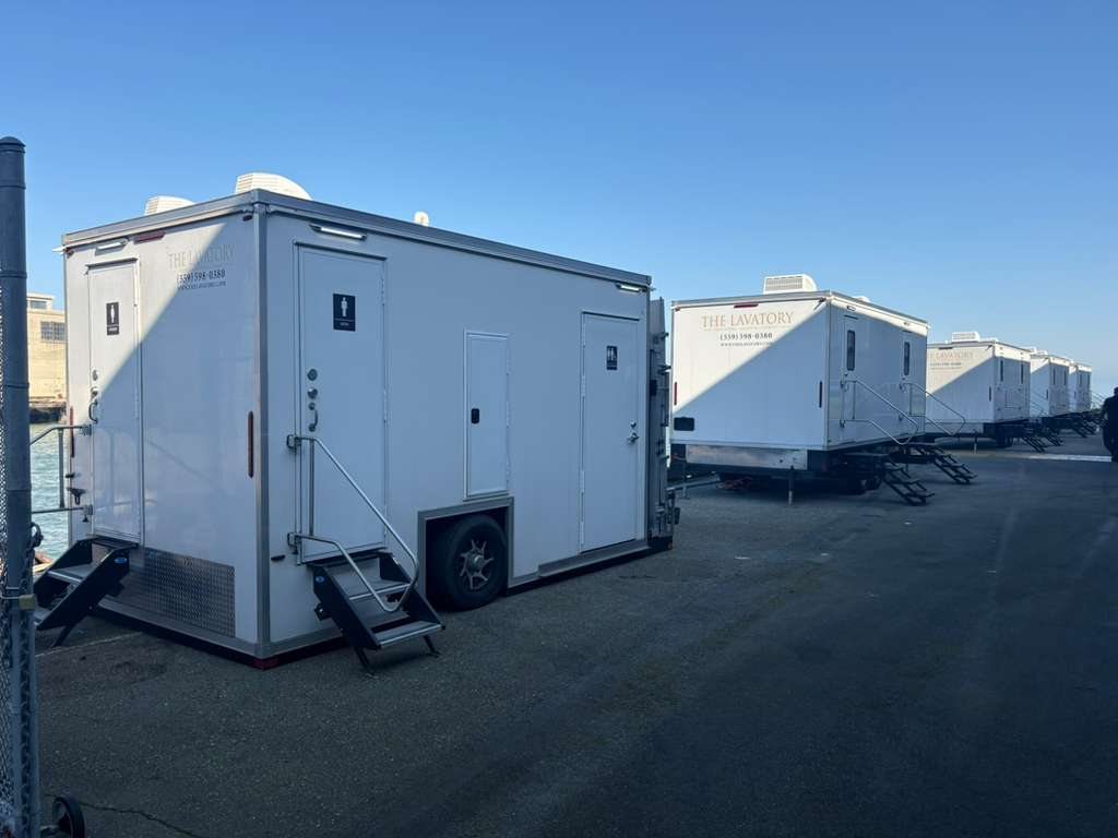 A row of Lavatory restroom trailers lined up on a paved waterfront lot, each with steps and handrails set at the doors