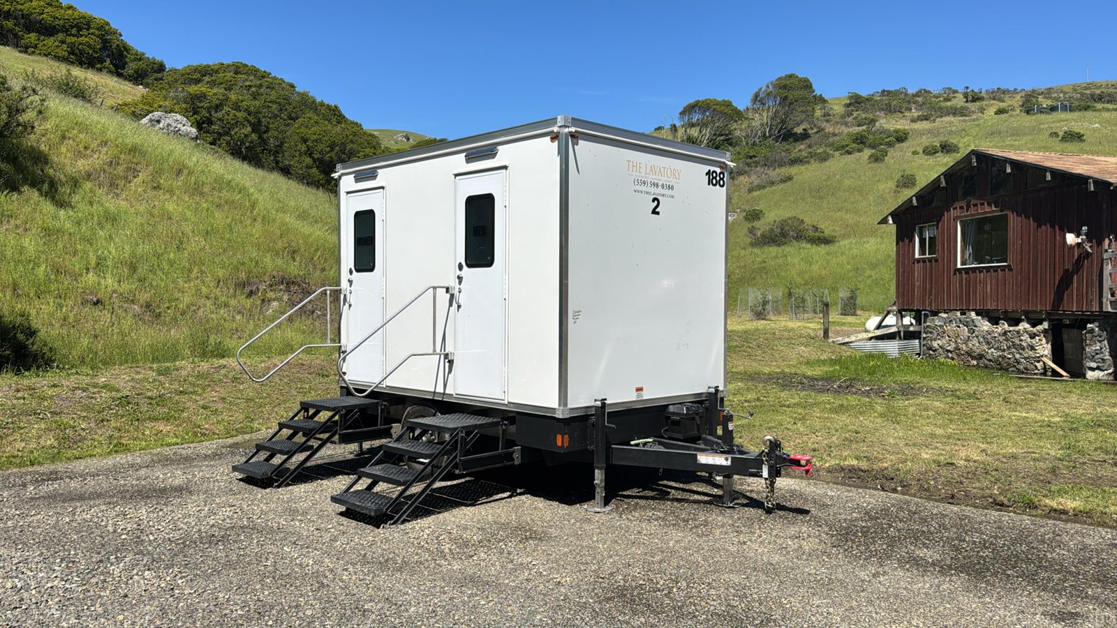 A two station Lavatory restroom trailer parked on gravel beside a wooden ranch building, with green hills behind it
