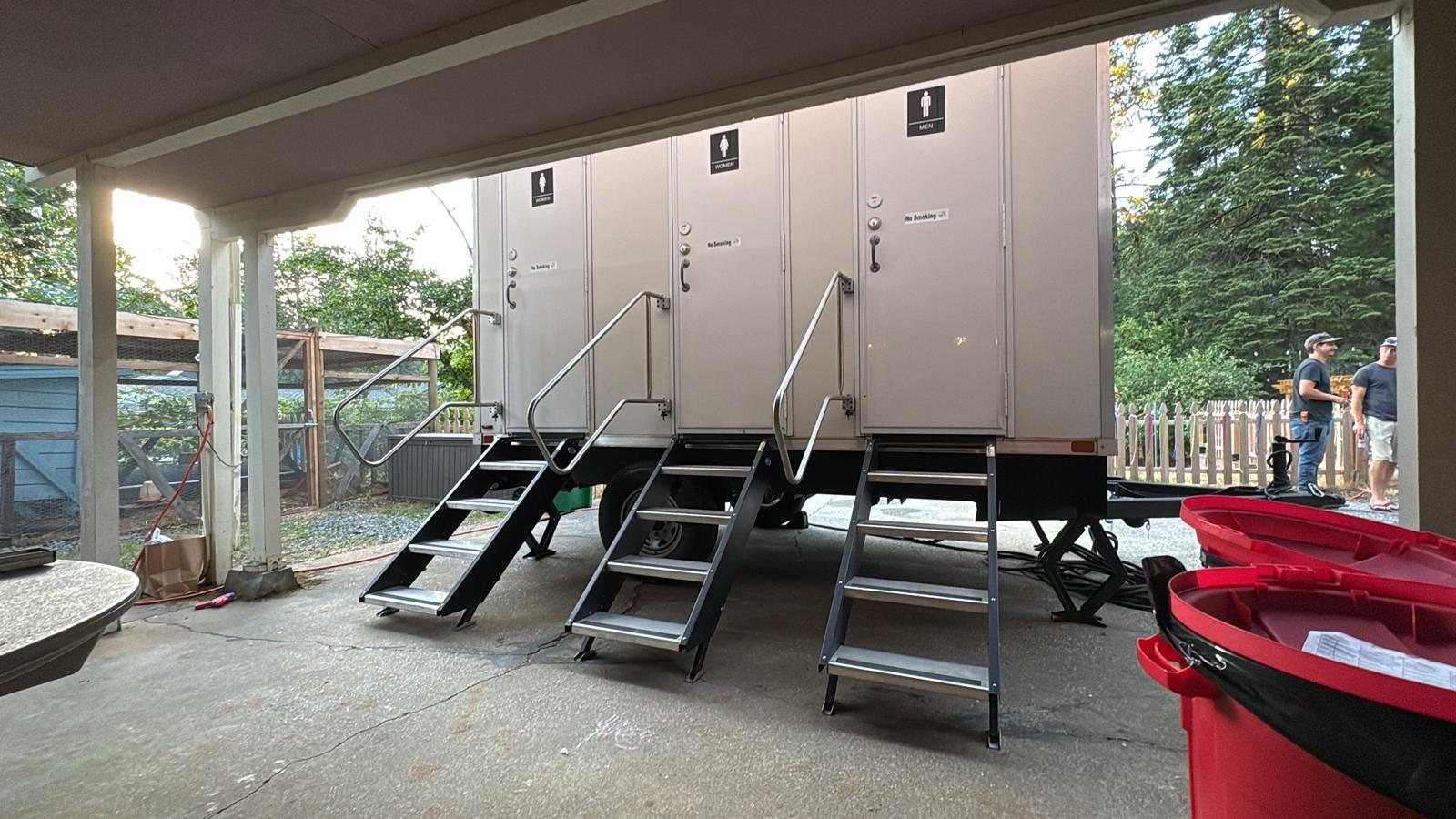 A three station Lavatory restroom trailer set under a covered patio at an outdoor event, with guests talking nearby