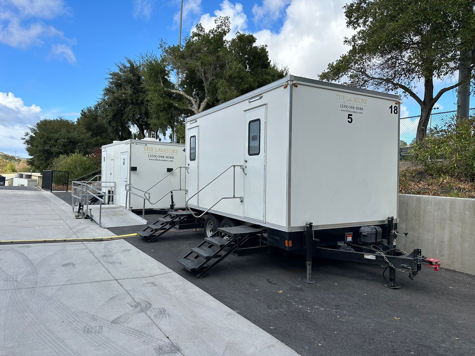 Two Lavatory restroom trailers set along a paved drive on a school campus, with steps at each door and trees behind them