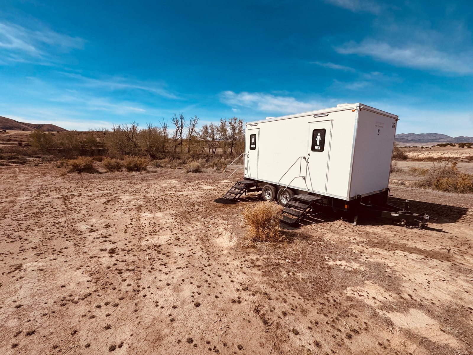 A Lavatory 5-Station restroom trailer set on open desert ground with dry brush and low mountains under a blue sky