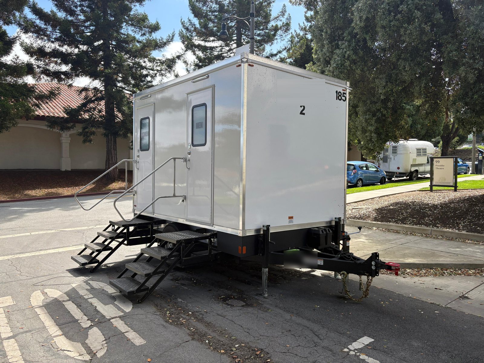 A Lavatory 2-Station restroom trailer on a paved lot beside tall redwood trees and a tile-roofed building
