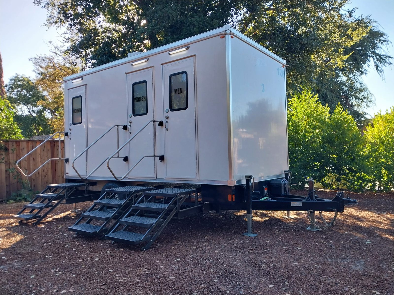 A Lavatory 3-Station restroom trailer on bark mulch in a fenced backyard under large oak trees
