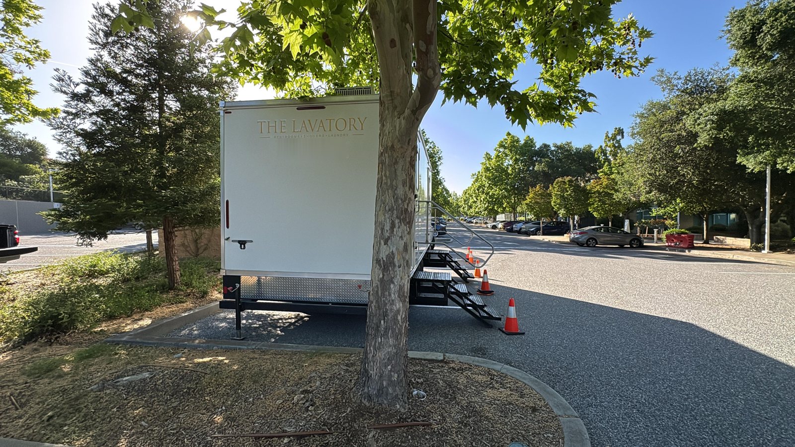 A Lavatory restroom trailer parked under a sycamore tree in an office parking lot in Palo Alto, with orange cones beside the steps