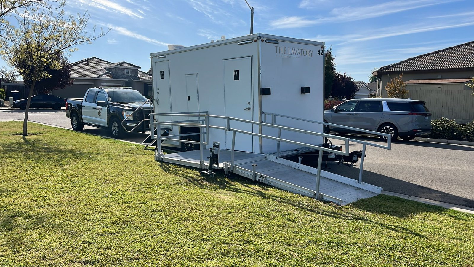 A Lavatory ADA restroom trailer with its aluminum ramp set on a grass strip beside a residential street
