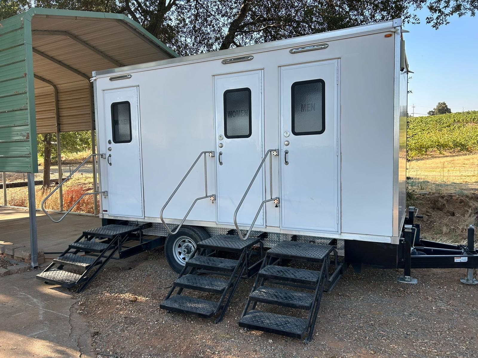 A Lavatory 3-Station restroom trailer on gravel beside a carport, with vineyard rows behind it