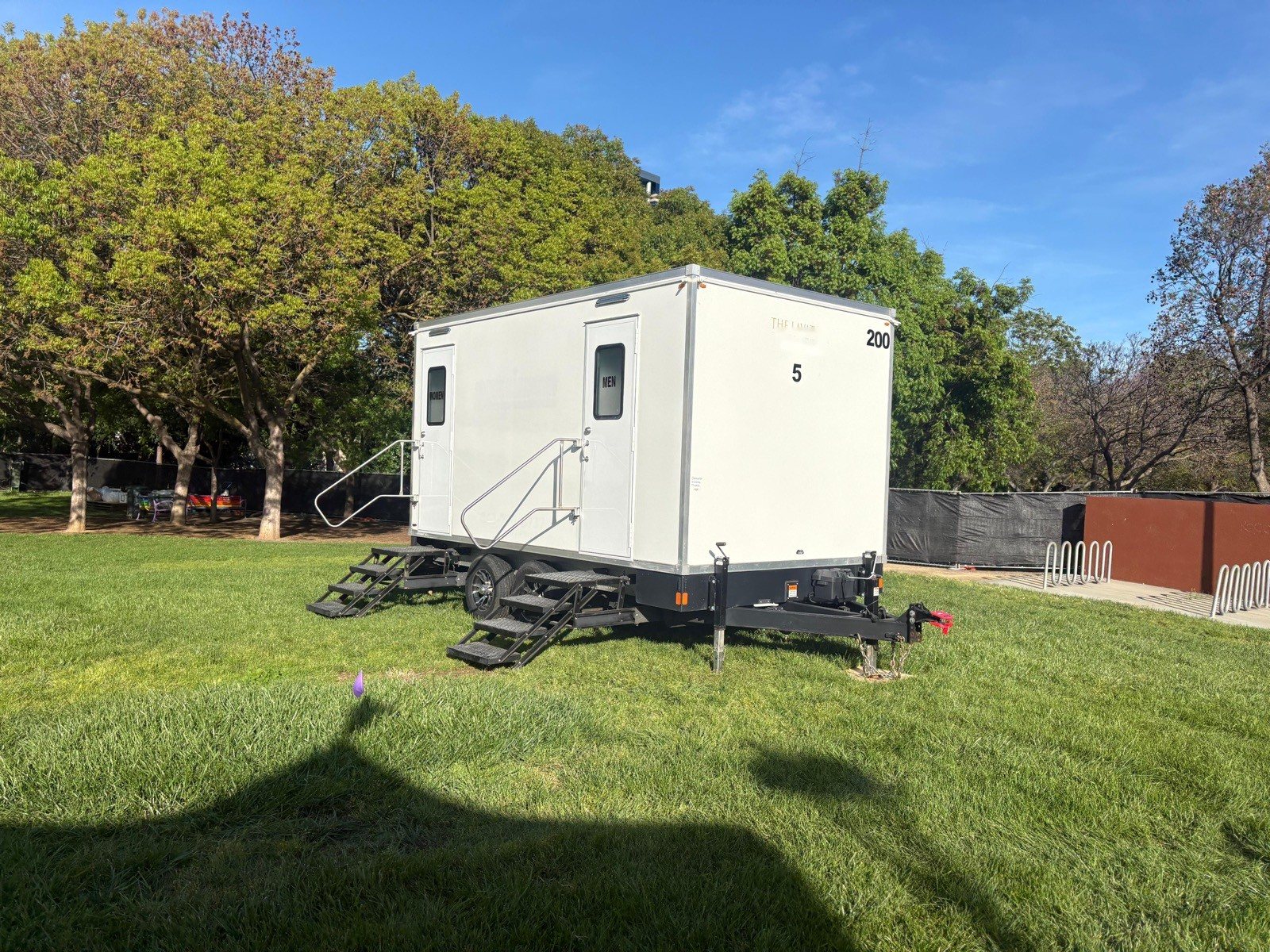 A Lavatory 5-Station restroom trailer on a green park lawn with shade trees behind it