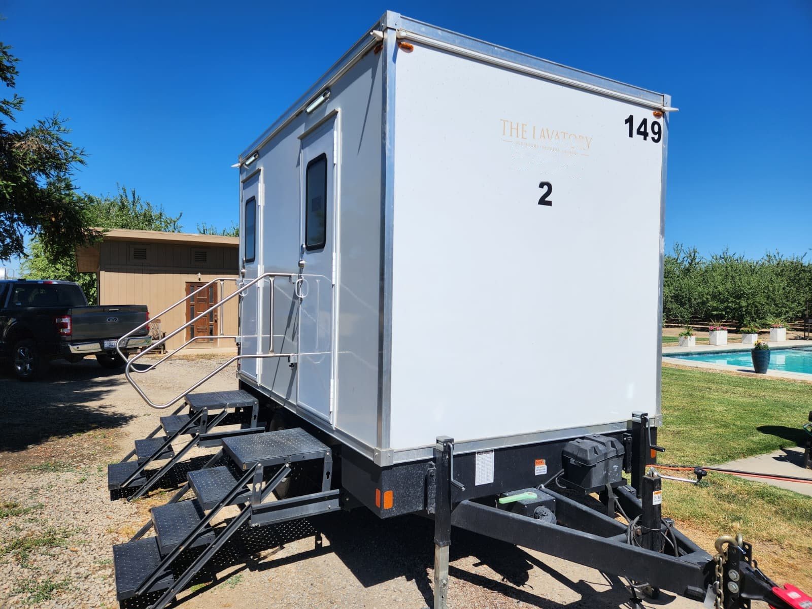 A Lavatory 2-Station restroom trailer on a gravel drive beside a swimming pool and a small pool house