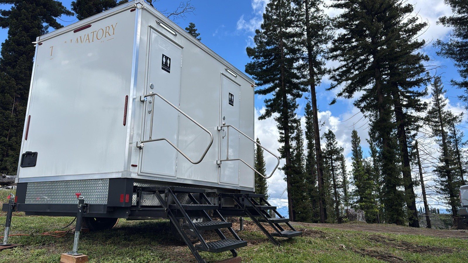 A Lavatory 2-Station restroom trailer set up on a grassy lot among tall pine trees
