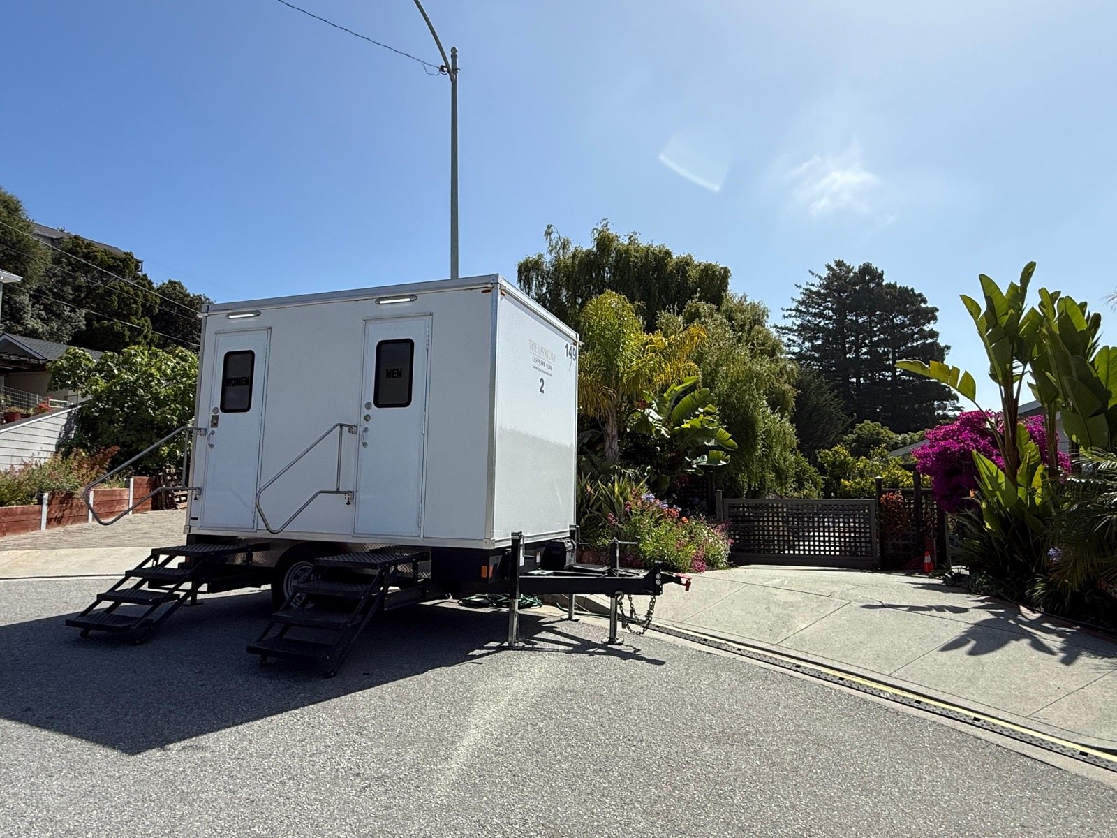 A Lavatory 2-Station restroom trailer parked at the curb of a residential street beside a flowering garden