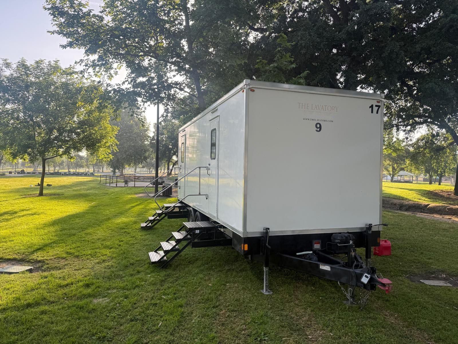 A Lavatory 9-Station restroom trailer parked on a park lawn in morning light, with shade trees behind it