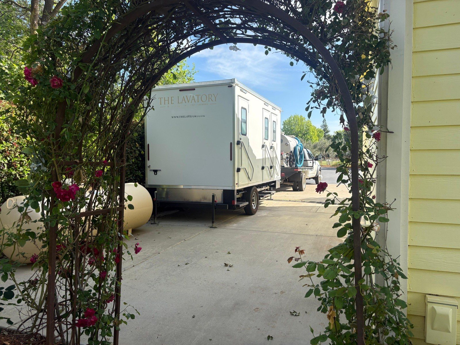 A Lavatory restroom trailer seen through a garden arch covered in climbing roses
