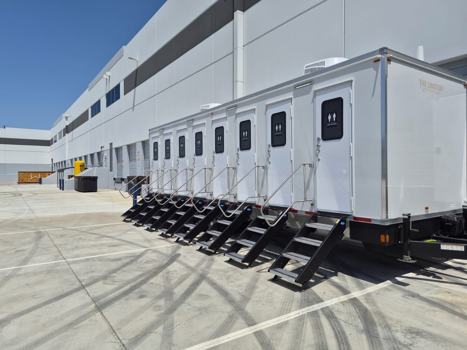 A long Lavatory 9-Station restroom trailer parked on a paved lot beside a large building, with stairs at every door