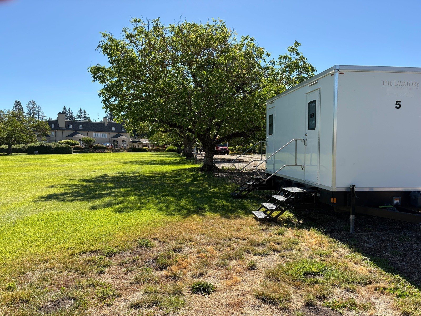 A Lavatory 5-Station restroom trailer on the edge of a wide lawn beside a spreading tree, with a large house in the distance