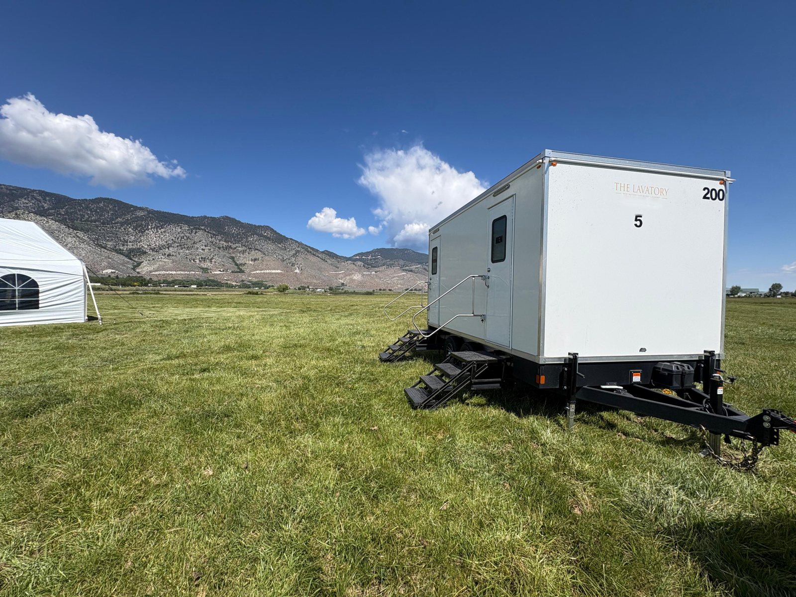A Lavatory 5-Station luxury restroom trailer on a green lawn beside a white event tent, with mountains behind it