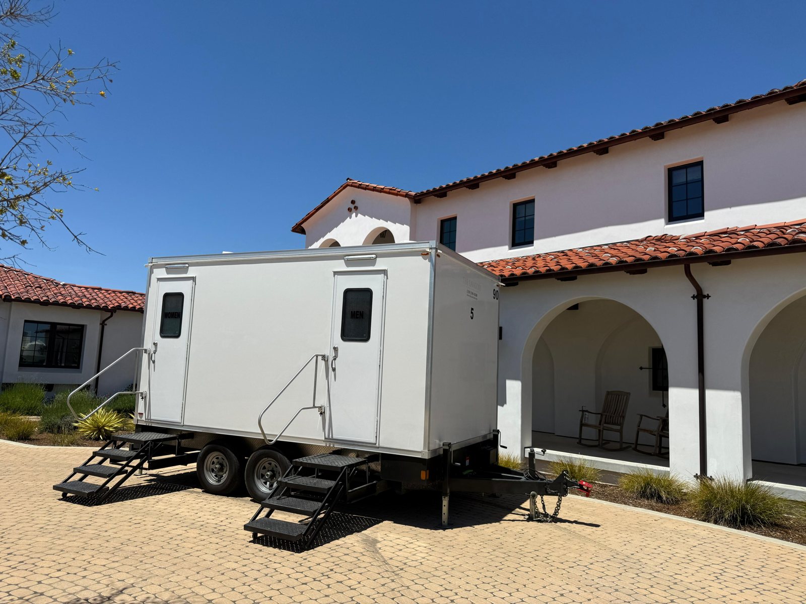 A Lavatory restroom trailer on a paver courtyard in front of a Spanish style building with red tile roofs and arches