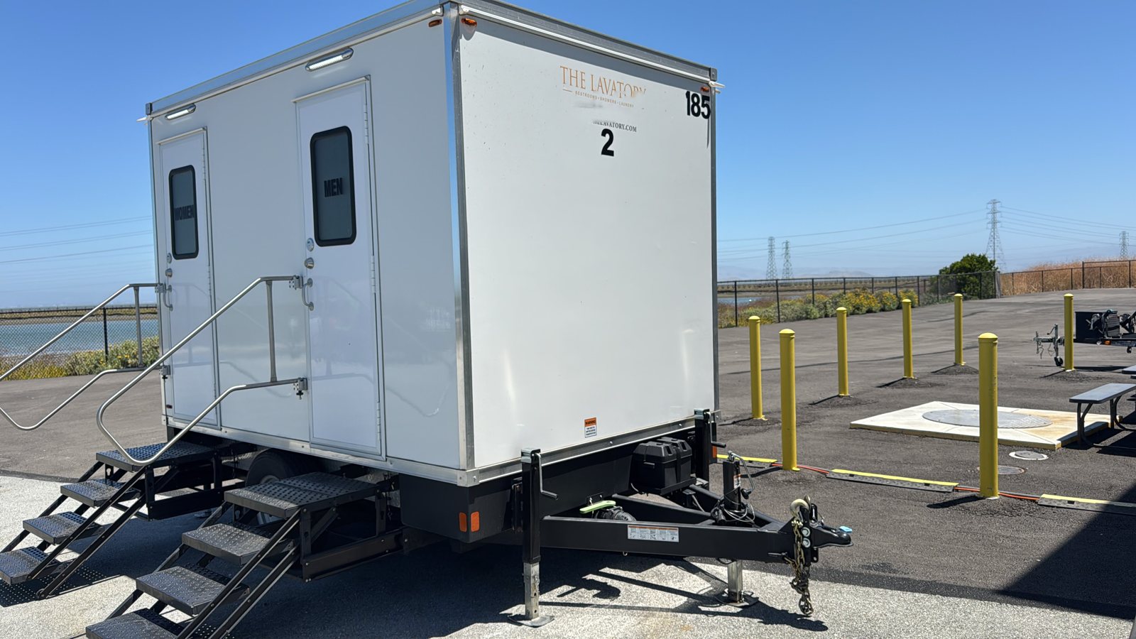 A Lavatory 2-Station restroom trailer parked on a paved lot with yellow bollards and open land behind it