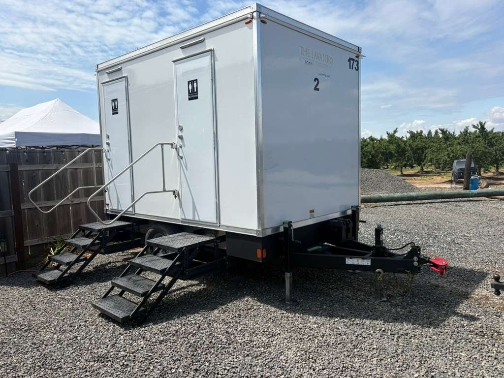A Lavatory 2-Station restroom trailer on a gravel lot beside an orchard, with a white event tent behind it