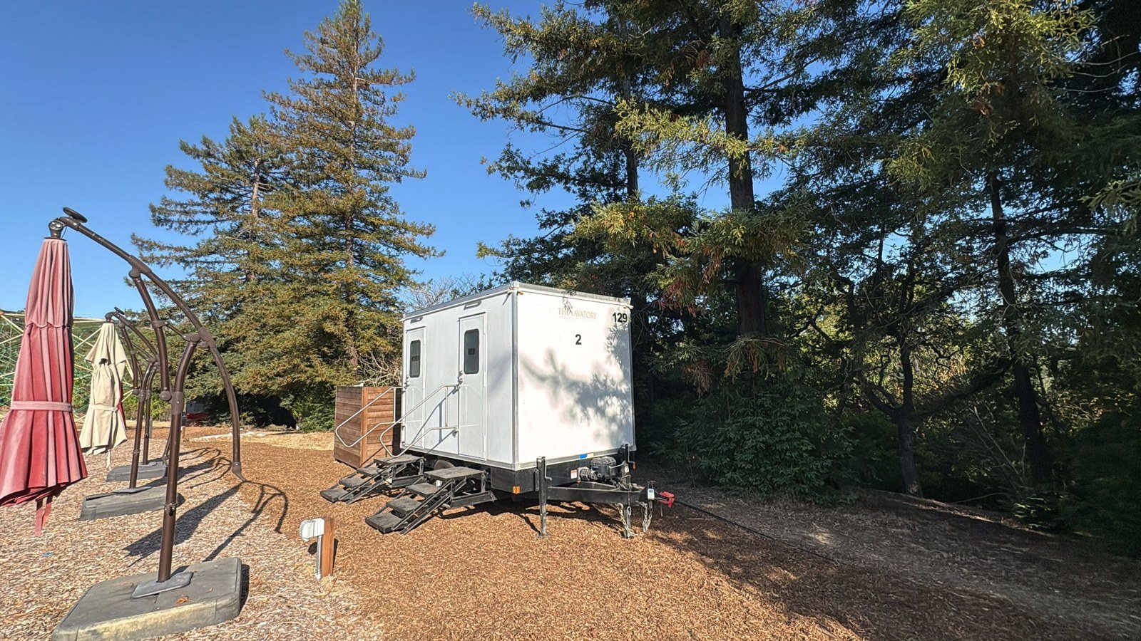 A Lavatory 2-Station restroom trailer on a wood-chip lot under tall evergreen trees in Redwood City, with patio umbrellas beside it