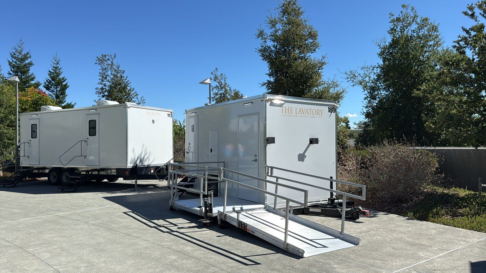 A Lavatory ADA + 2 restroom trailer with its aluminum ramp unfolded beside a 9-Station trailer on a corporate campus