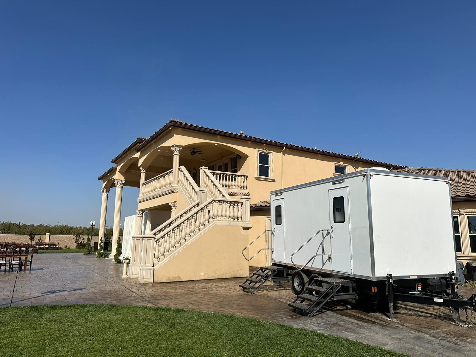 A Lavatory restroom trailer on a stamped concrete patio beside a villa-style wedding venue with a balustrade staircase