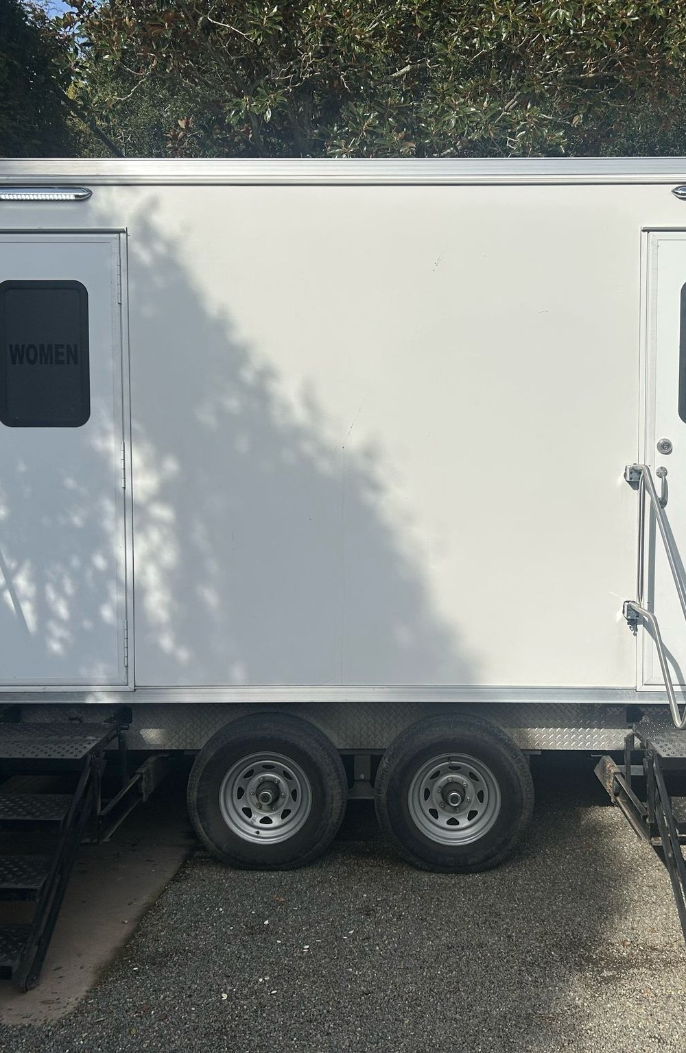 A Lavatory 2-Station restroom trailer with women's and men's doors on a gravel driveway beside a garage under oak trees