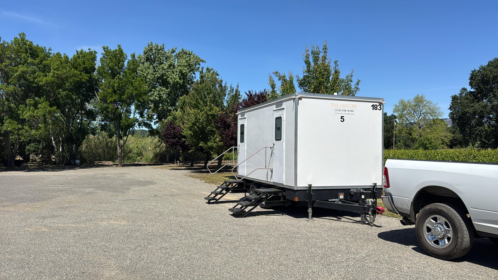 A Lavatory 5-Station restroom trailer on a gravel lot beside shade trees and a vineyard