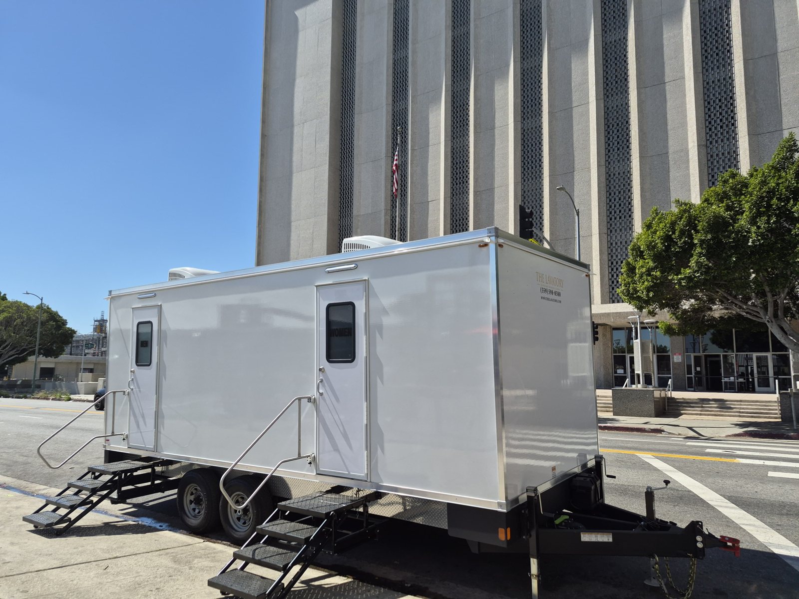 A Lavatory two-door restroom trailer parked curbside with its stairs down beside a tall civic building