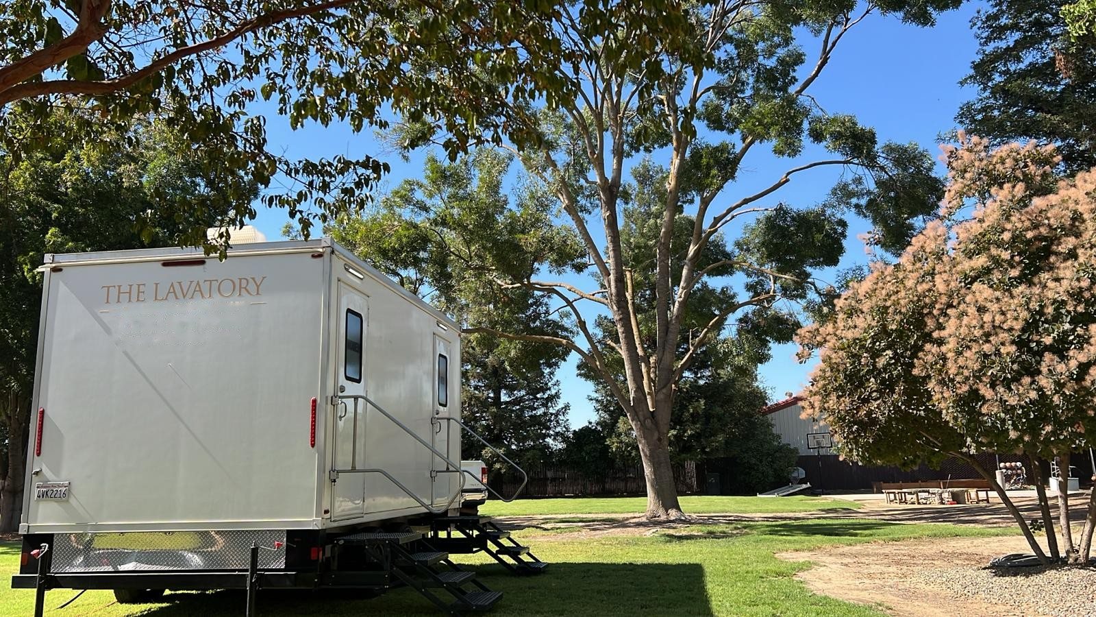 A Lavatory restroom trailer parked on a shaded lawn under tall trees beside a flowering shrub