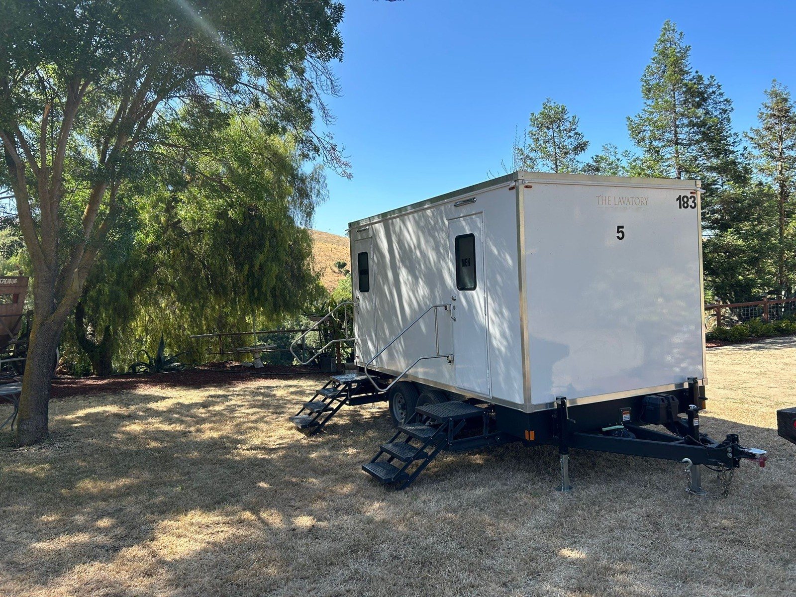 A Lavatory 5-Station restroom trailer on dry grass under a willow tree, with golden hills and pine trees behind it
