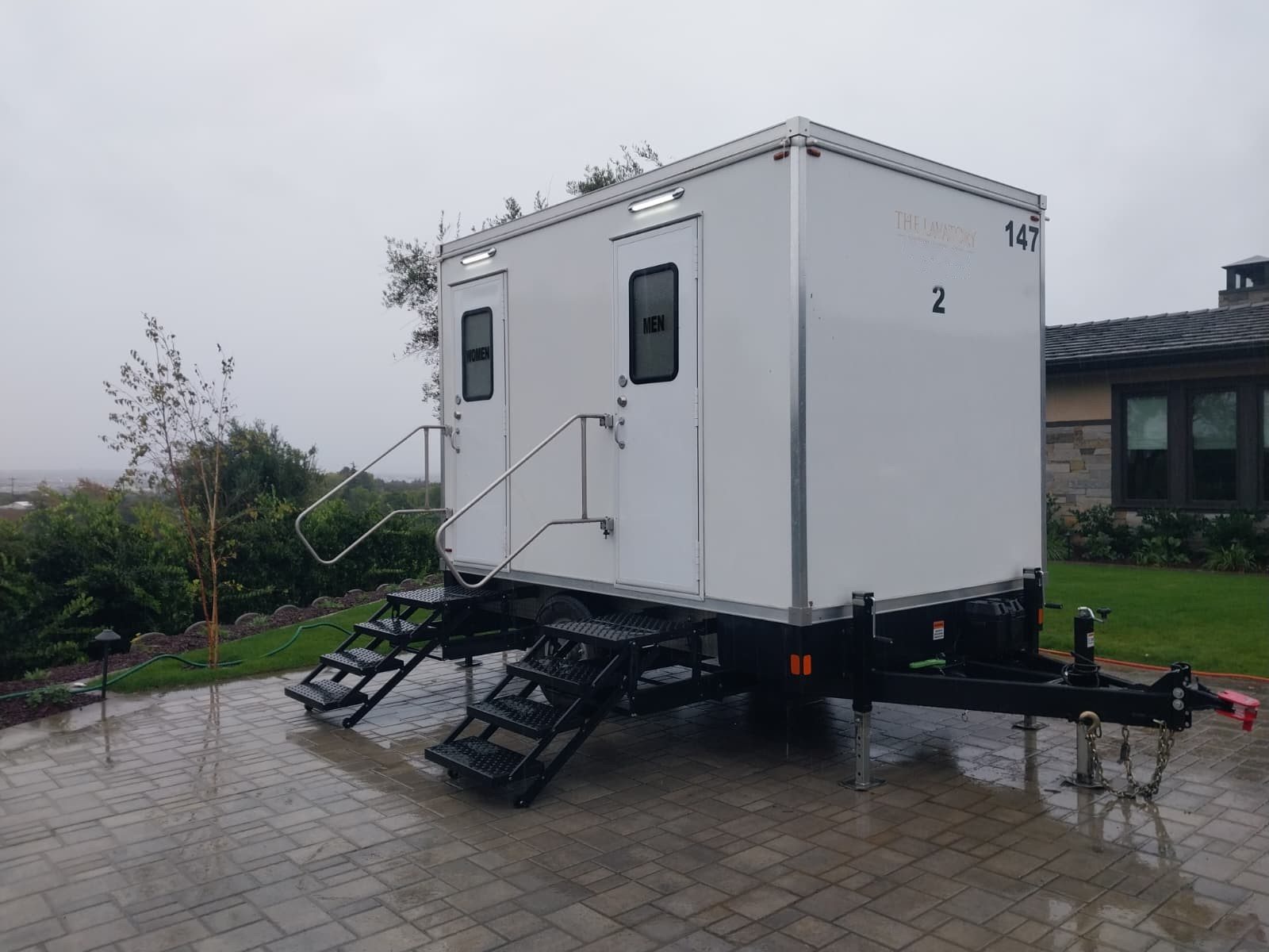 A Lavatory 2-Station restroom trailer on a paver patio beside a house and garden plants