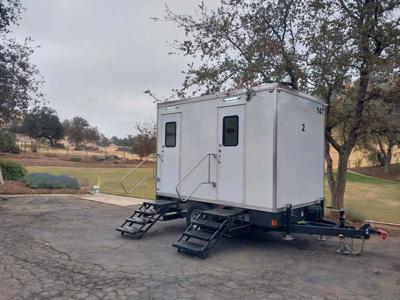 A Lavatory 2-Station restroom trailer on a paved pad under oak trees beside a green lawn
