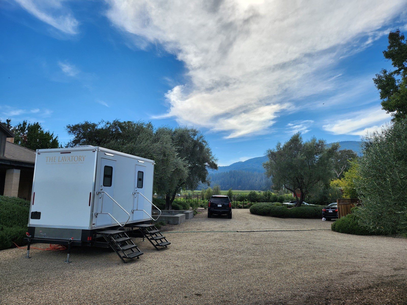 A Lavatory 2-Station restroom trailer on a gravel drive at a vineyard estate, with olive trees and hills behind it