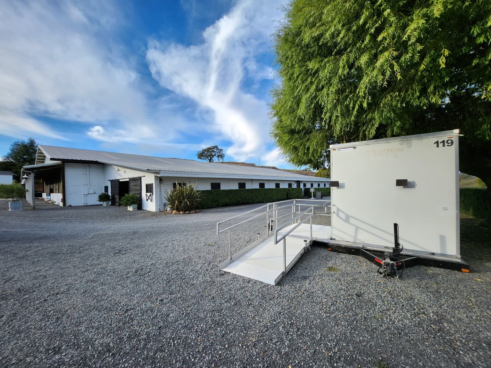 A Lavatory ADA restroom trailer with a metal ramp on a gravel yard next to a long white barn