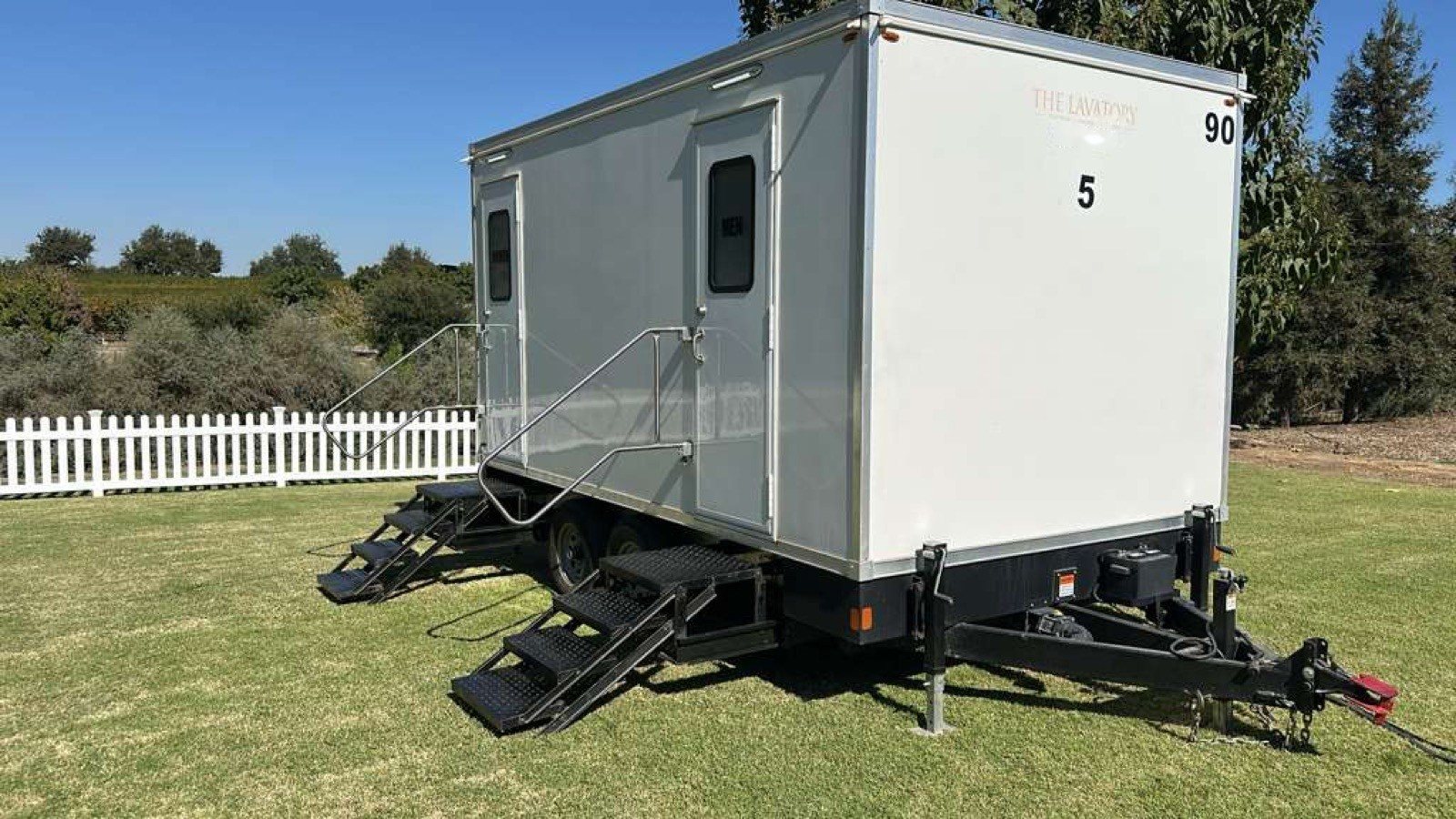 A Lavatory 5-Station restroom trailer on a lawn beside a white picket fence