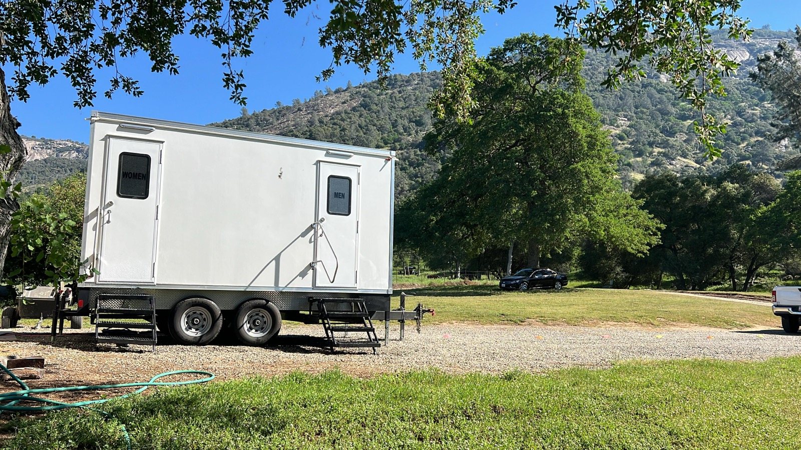 A Lavatory restroom trailer on a lawn at a rural property with green hills and oak trees behind it