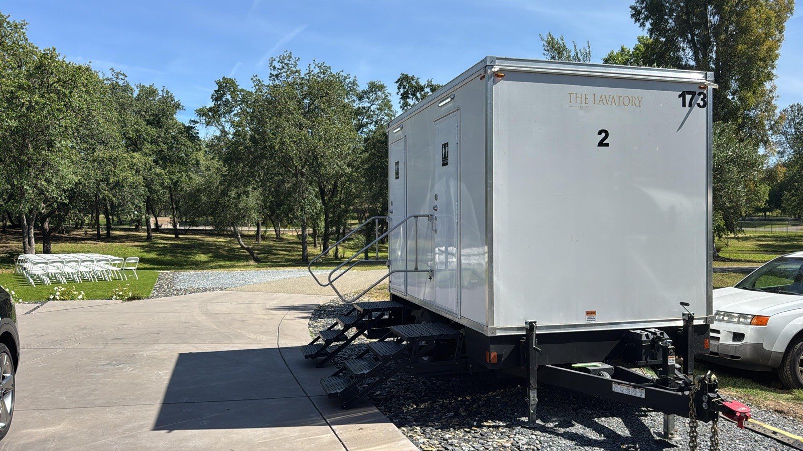 A Lavatory 2-Station restroom trailer parked on a gravel drive in an oak grove, with white chairs set on the lawn