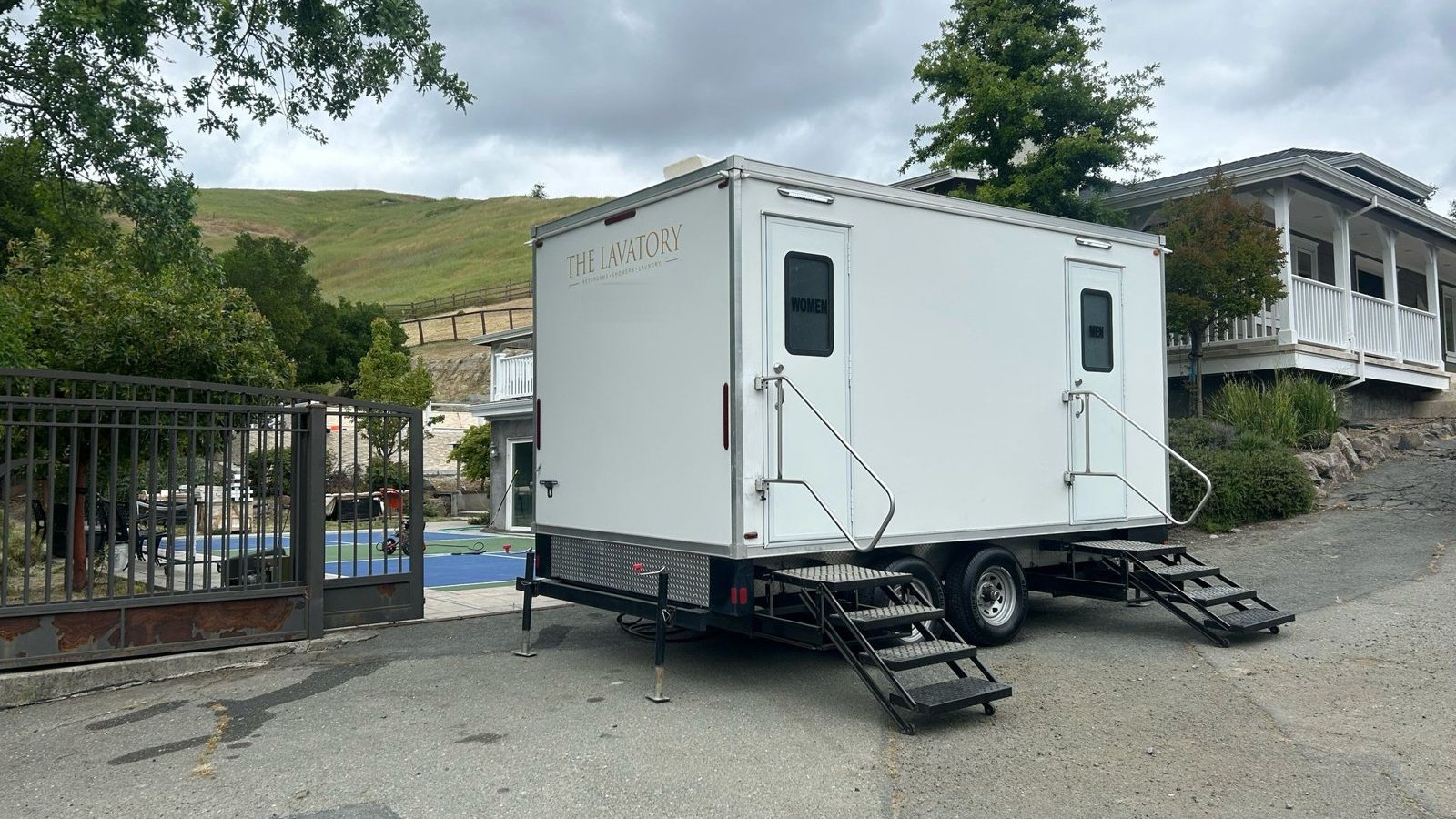A Lavatory 2-Station luxury restroom trailer with steel stairs and handrails parked on the driveway of a hillside home, beside an iron gate and a backyard sport court