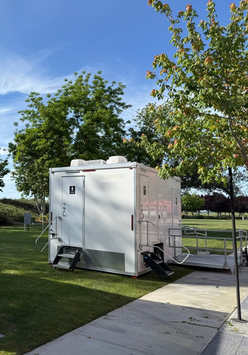 A Lavatory restroom trailer with an aluminum ADA ramp and landing on a park lawn beside a paved walkway under shade trees