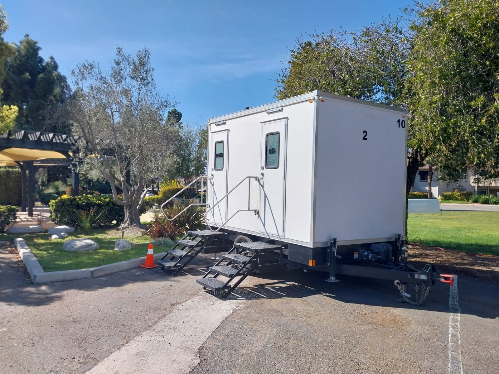 A Lavatory 2-Station restroom trailer parked beside a landscaped garden with an olive tree and a shade pergola