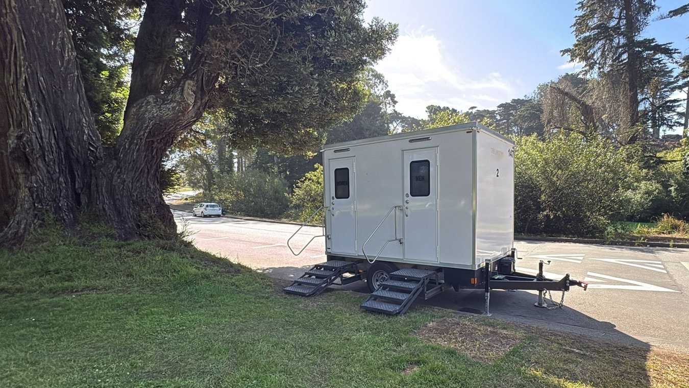 A Lavatory 2-Station restroom trailer parked on the grass edge of a park road under large cypress trees