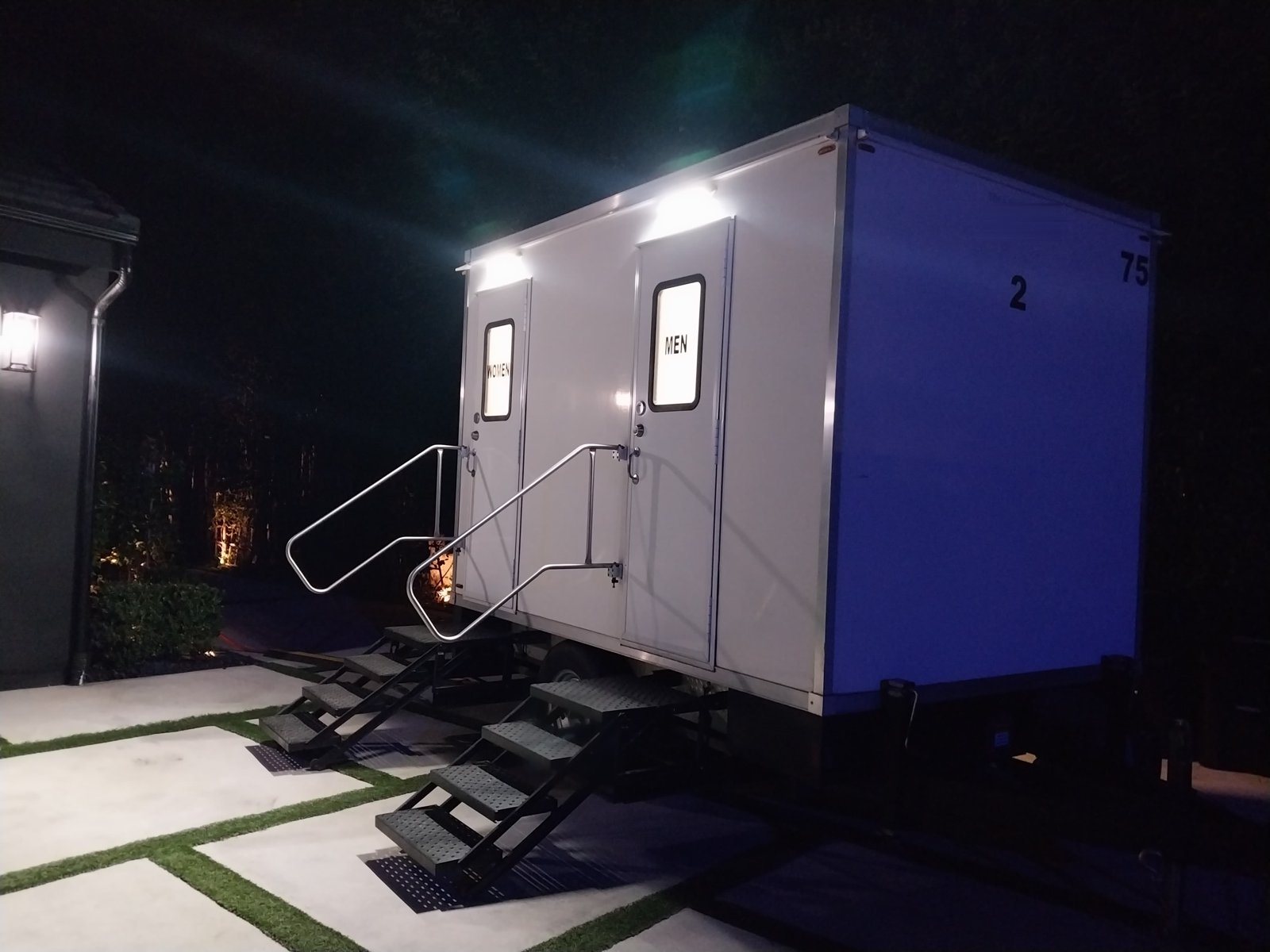 A Lavatory 2-Station restroom trailer lit up at night on a paver driveway, with lights above both doors and lit stair rails