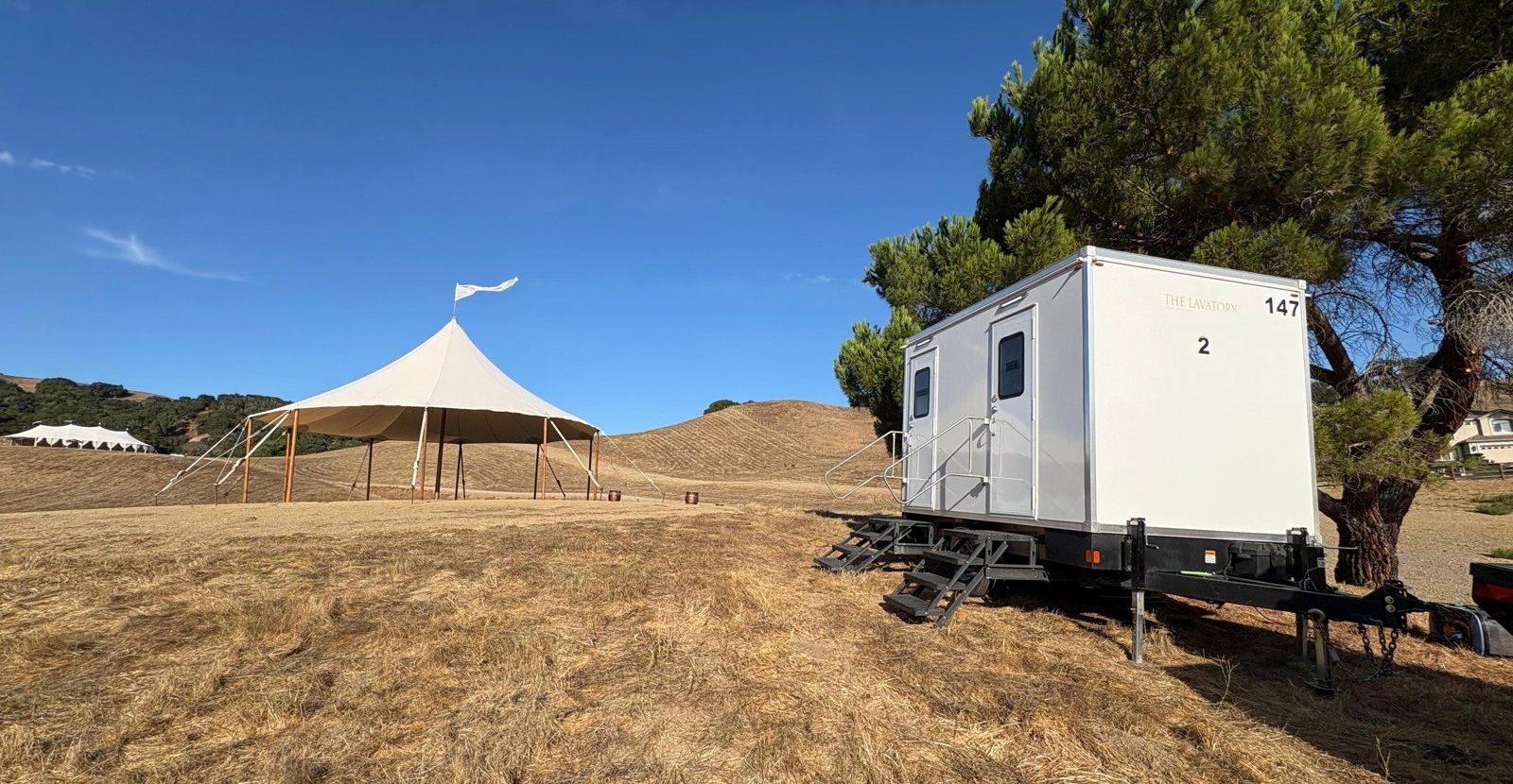 A Lavatory 2-Station luxury restroom trailer under a pine tree on golden grass hills, with a sailcloth wedding tent on the field beside it