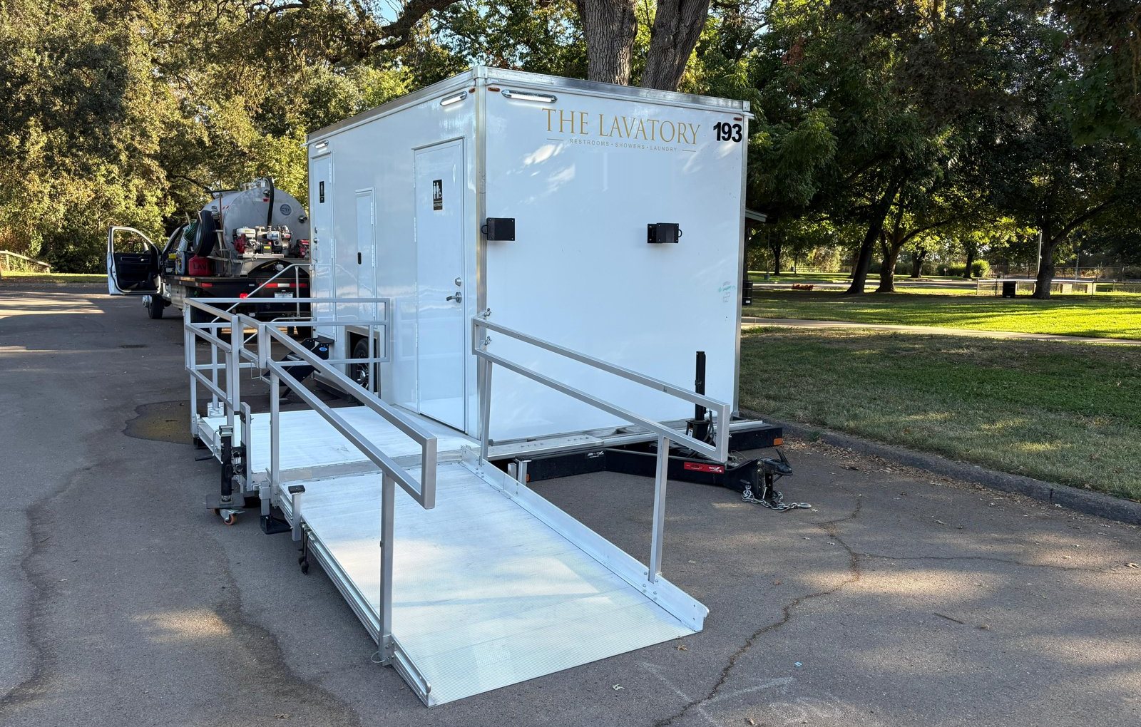 A Lavatory ADA restroom trailer with a wheelchair ramp in a park parking lot under shade trees, with a service pump truck behind it